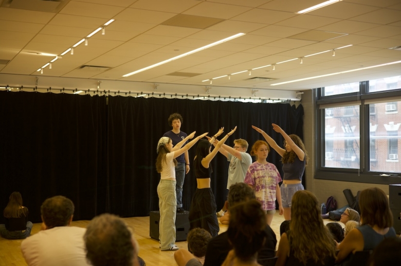 A group of Tisch Summer HIgh School Drama students stand on a stage during their final presentations on the last day of class. Behind them is a black curtain. Two groups of two students face each other with arms raised at an angle, forming a tunnel as one student walks through. Another student stands on a raised box behind the students forming the tunnel.